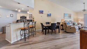 A modern kitchen with a central island, dining area with chairs, and a living room with a sofa and lamp.