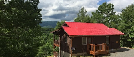 A cabin with a red roof, a wooden deck, and a view of the surrounding forest and mountains.