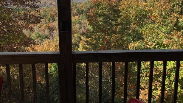 Une terrasse en bois avec une table et une chaise rouge, d’où l’on peut admirer un paysage boisé aux couleurs automnales et des montagnes lointaines.