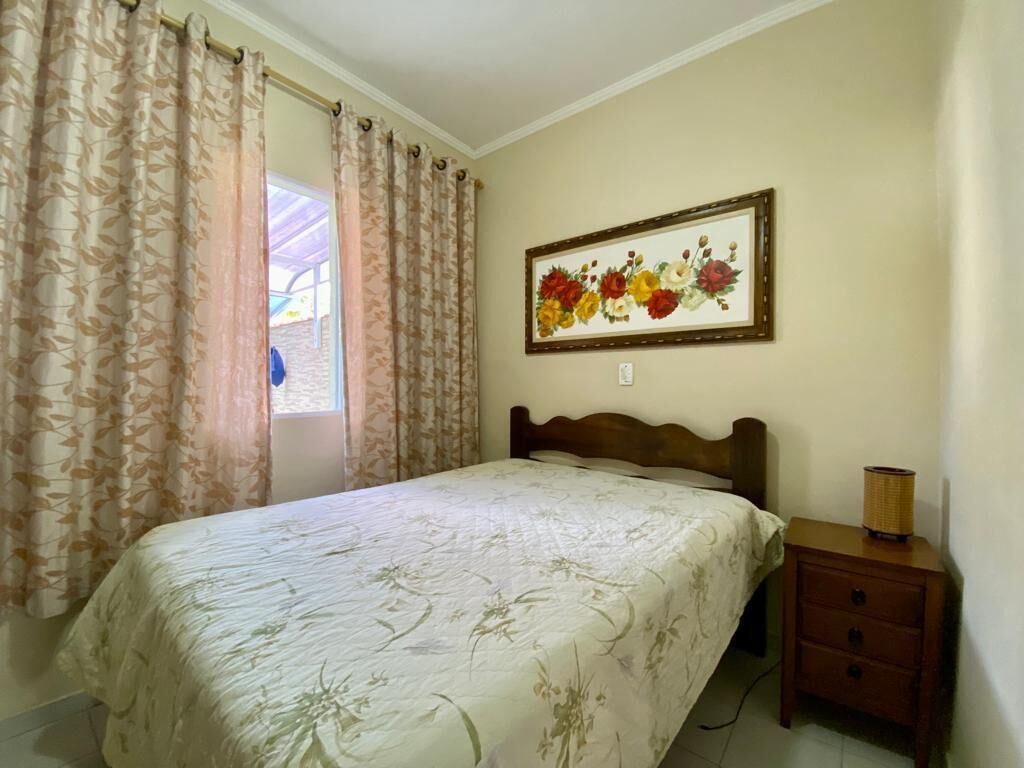 Quarto com cama, mesa de cabeceira, luminária e um quadro com flores na parede.