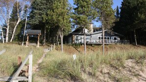 A cabin with a deck and a covered porch, surrounded by tall pine trees and a sandy path leading to it. - 8 Miles Of Sand Beach To Explore ~ Outstanding Sunrise Views Over The Lake! (Sturgeon Bay)