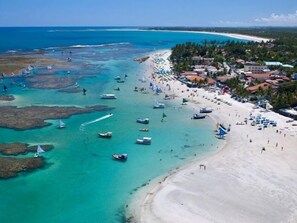 An aerial view of a beach with boats, a sandy shore, and buildings in the background.