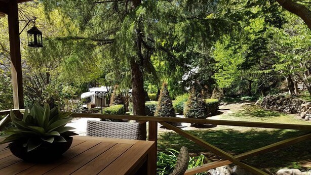 Una terrassa de fusta amb taula i cadires, que dona a un jardí ple de plantes diverses i una petita bassa.