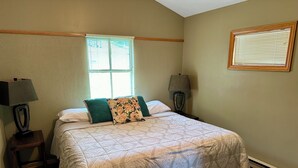 A bedroom with a bed, two bedside tables, a window with blinds, and a wall-mounted lamp.
