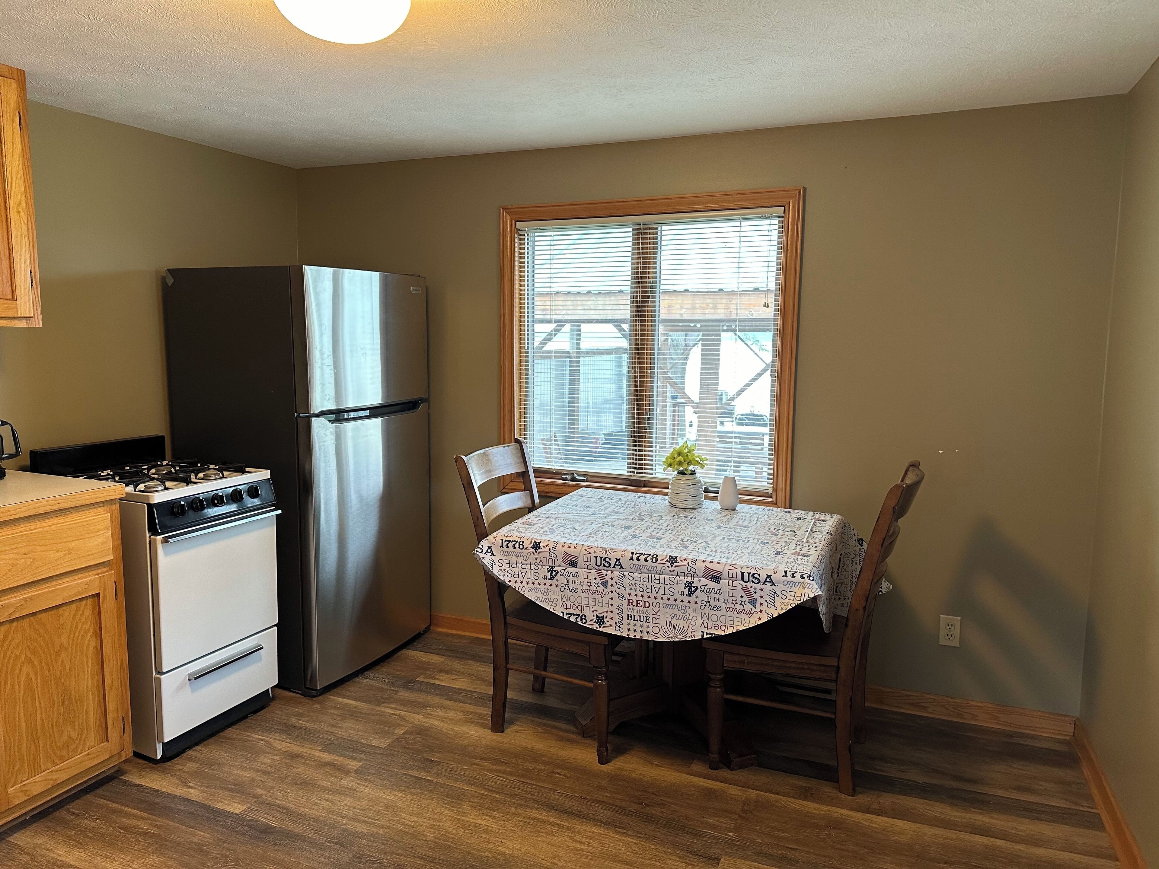 Une cuisine compacte équipée d’une cuisinière, d’un four et d’un réfrigérateur. Une table à manger avec des chaises est placée près d’une fenêtre munie de stores.
