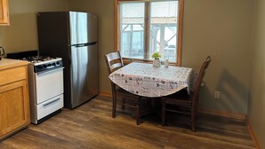 A compact kitchen with a stove, oven, and refrigerator. A dining table with chairs is positioned near a window with blinds.