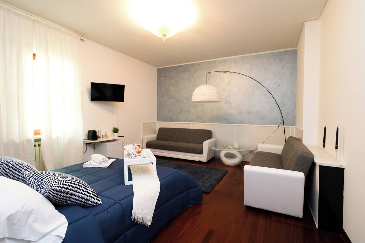 A modern hotel room with a bed, a sofa, a TV, a lamp, and a small table.