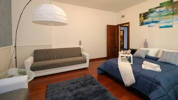 Ein modernes Wohnzimmer mit grauem Sofa, blauem Sofa, Glastisch, Stehlampe und Kunstwerken an der Wand.