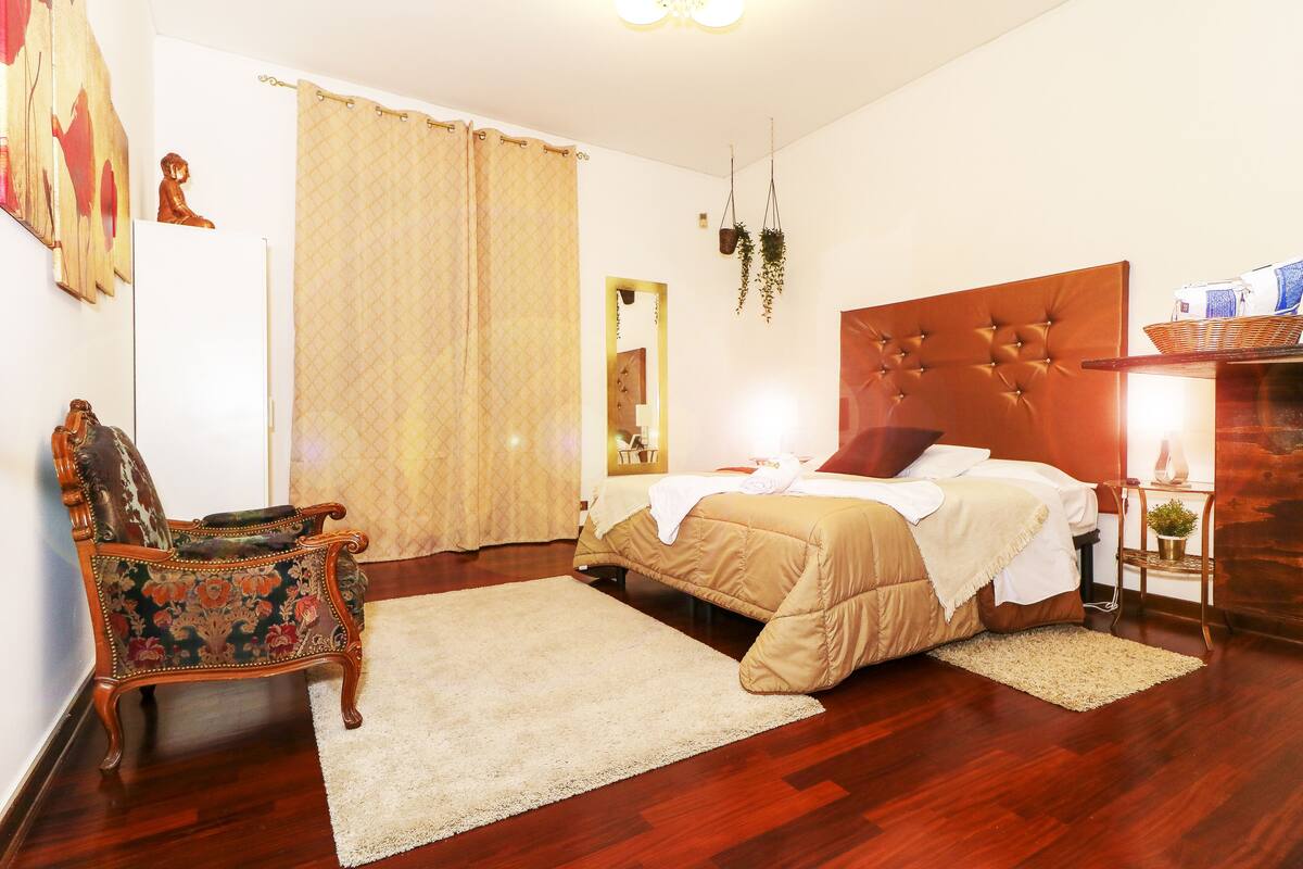 A bedroom with a wooden headboard bed, a decorative chair, a large mirror, and a wooden nightstand with a lamp.