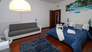 Ein modernes Schlafzimmer mit einem grauen Sofa, einem blauen Bett mit weißer Decke, Holzboden und einem an der Wand befestigten Kunstwerk, das einen Wasserfall zeigt.