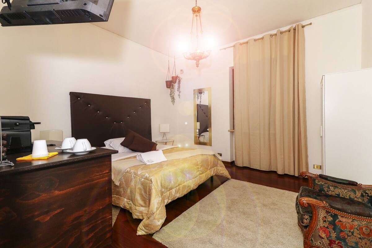 A hotel room with a bed, a desk with a lamp, a chair, a mirror, and a window with curtains.