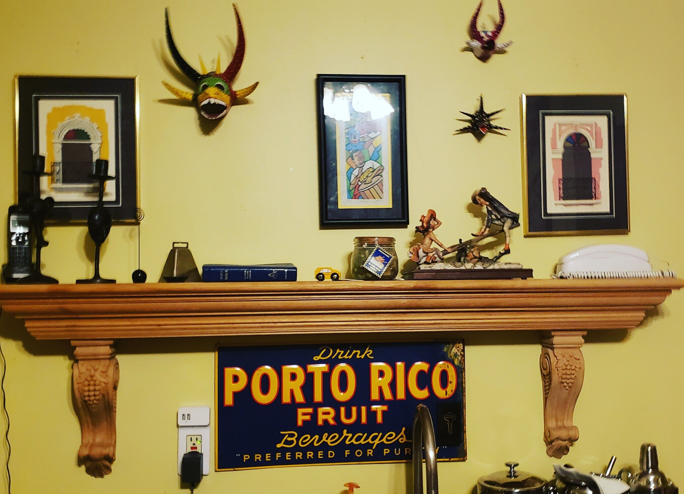 Una habitación con una repisa de madera que exhibe varios objetos, un letrero enmarcado de 'Porto Rico Fruit Beverages' y adornos de pared que incluyen un cráneo y una fotografía enmarcada.