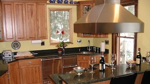 Una cocina con armarios de madera, campana extractora de acero inoxidable, un ventanal grande y una zona de bar con una botella de vino y copas.