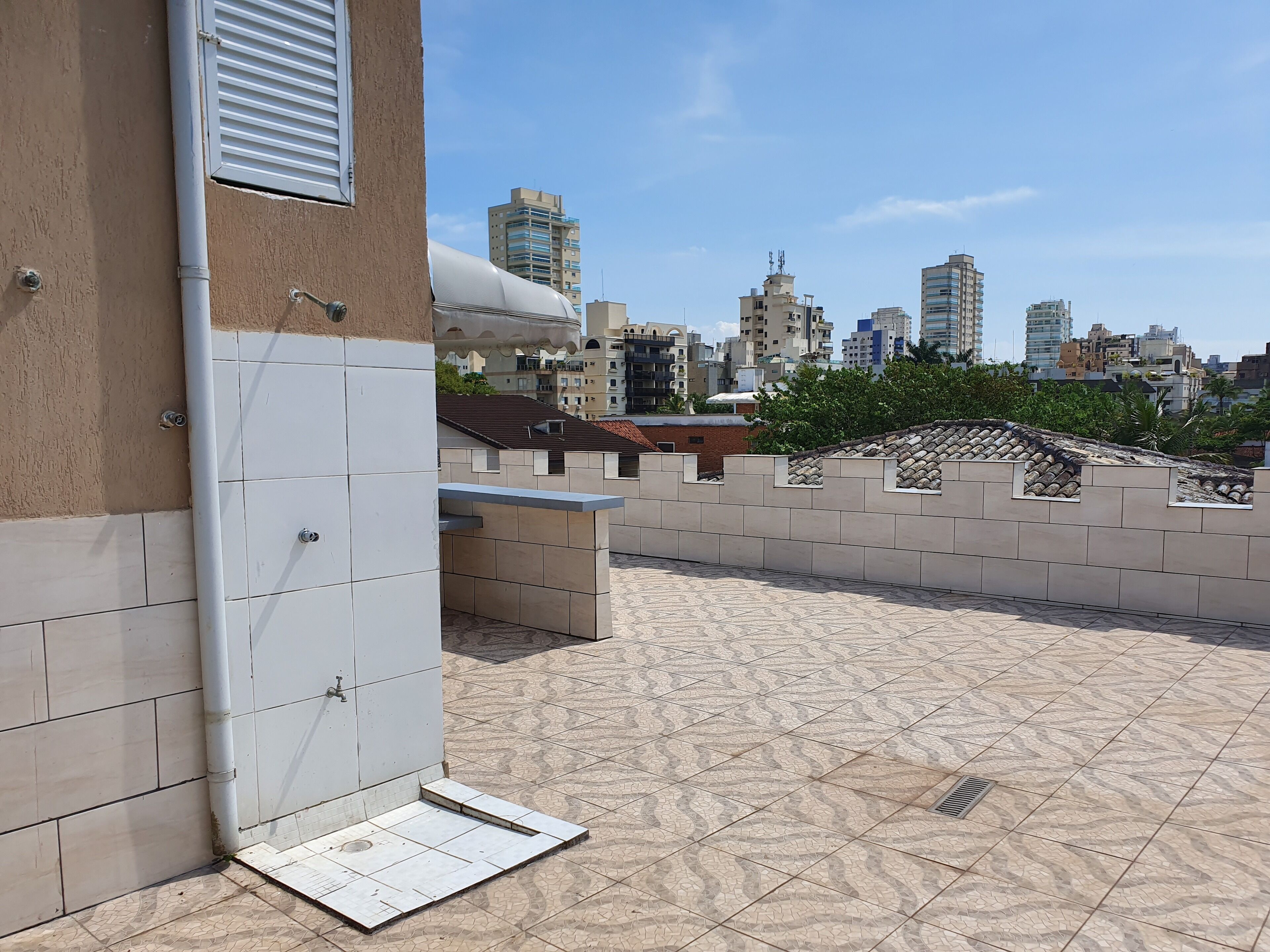 Een terras op het dak met betegelde vloer, een muur met een raam en een uitzicht op een stadsbeeld met gebouwen op de achtergrond.