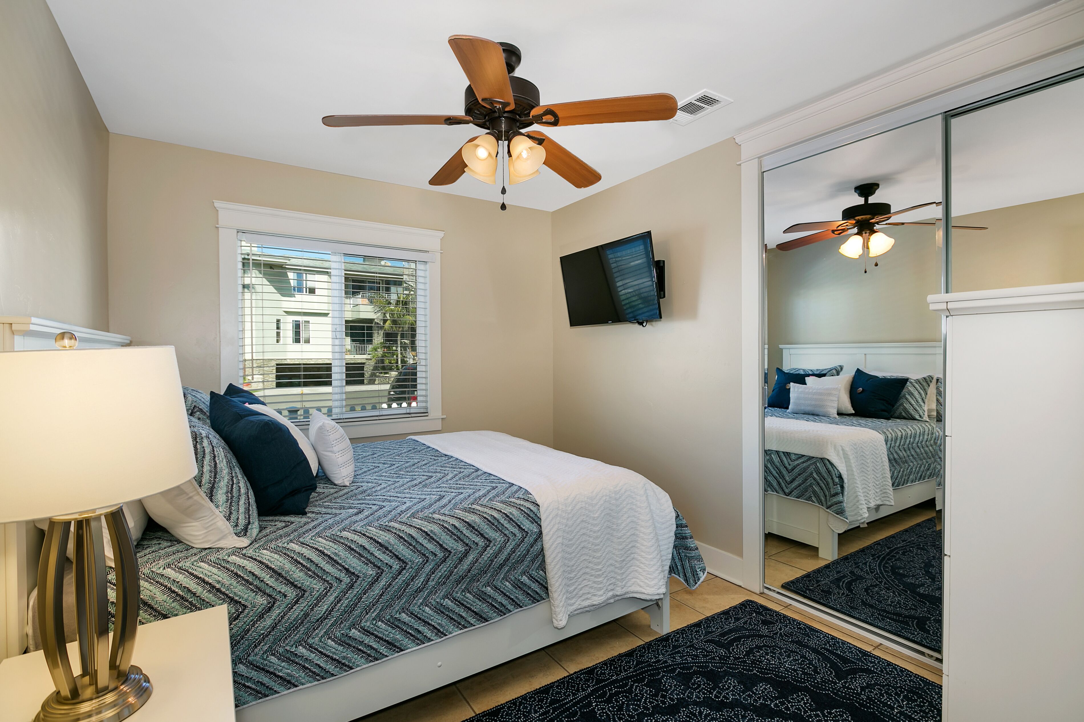 A bedroom with a bed, a ceiling fan, a window, a TV, and a mirror.