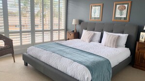 A bedroom with a large bed, a nightstand, a lamp, and a chair. There are two framed botanical prints on the wall. - Barefoot, 7/44 Marine Drive - Fabulous Fingal Bay (Fingal Bay)