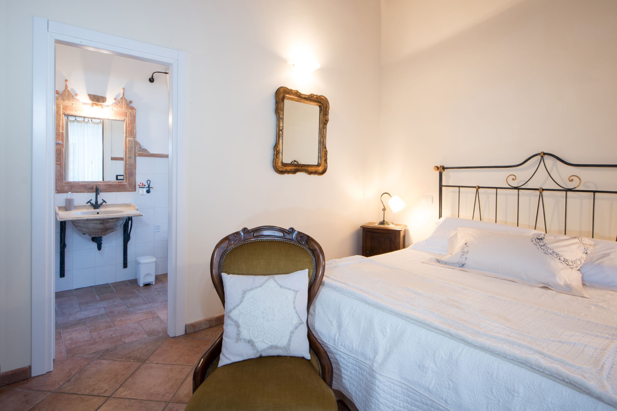 Quarto com cama, poltrona, mesa de cabeceira, espelho e banheiro com pia e vaso sanitário.