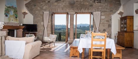 A rustic room with a stone wall, a fireplace, a TV, a dining table with chairs, and a view of the countryside.
