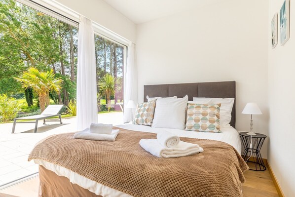 A bedroom with a large bed, a view of the outdoors, and a small table with a lamp.