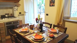 Une salle à manger avec une table en bois dressée pour quatre personnes, entourée de chaises. On aperçoit une cuisine à l’arrière-plan, avec une cuisinière et un four.