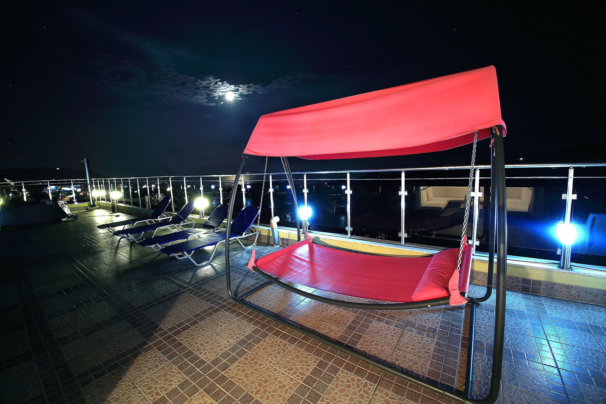 A red canopy swing chair on a balcony with tiled flooring and a railing.