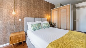 Ein Schlafzimmer mit Wand im Fischgrätmuster, einem Bett mit weißer Bettwäsche, zwei grün gemusterten Kissen, einem Nachttisch aus Holz und einer gelben Tagesdecke.