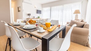 Moderni ruokailutila, jossa on puinen pöytä, valkoiset tuolit sekä aamiaistarvikkeita, kuten croissantteja ja appelsiinimehua.