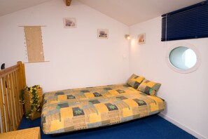 A small, single-bed room with a blue carpet, a wooden headboard, a round window, and a wall-mounted shelf with decorative items. - Maison cosy à 250m du lac | Entrée privée! (St Jean de Monts)