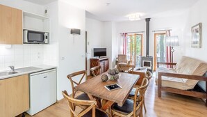 Una cocina moderna y compacta con armarios blancos, microondas y lavamanos. Hay un comedor con mesa y sillas de madera, una televisión y una chimenea. La habitación tiene ventanas grandes y un sofá.