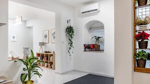 Un interior moderno con una planta grande en maceta, una alfombra gris y una estantería llena de libros y objetos decorativos.