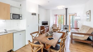 A compact, modern kitchen with white cabinets, a microwave, and a sink. There is a dining area with a wooden table and chairs, a television, and a fireplace. The room has large windows and a sofa.
