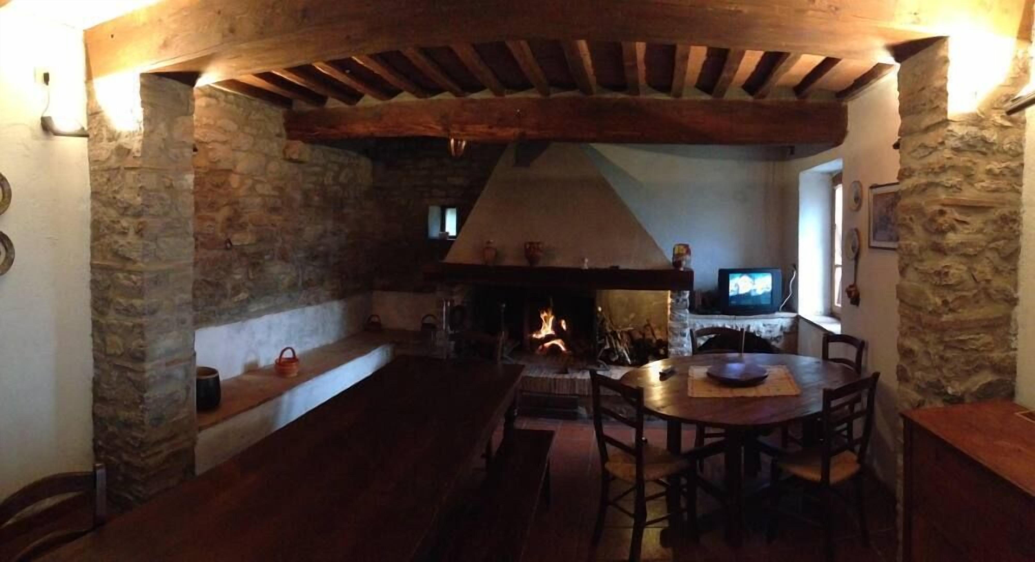 Um interior rústico com vigas de madeira aparentes, paredes de pedra, lareira e uma mesa redonda com cadeiras.