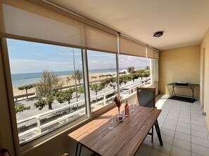 Habitació amb un gran finestral que dona a una platja, una taula de fusta amb copes de vi i una taula petita amb una bossa.