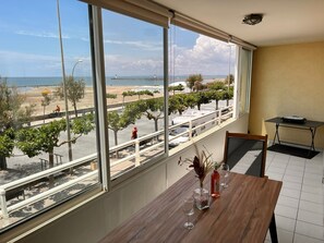 Ein Zimmer mit großem Fenster mit Blick auf einen Strand, ein Holztisch mit einer Vase und Weingläsern sowie ein kleiner Tisch mit einer Flasche.