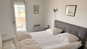 Un letto rifatto con lenzuola e cuscini bianchi, una testiera grigia rivestita in tessuto e due quadri appesi al muro.