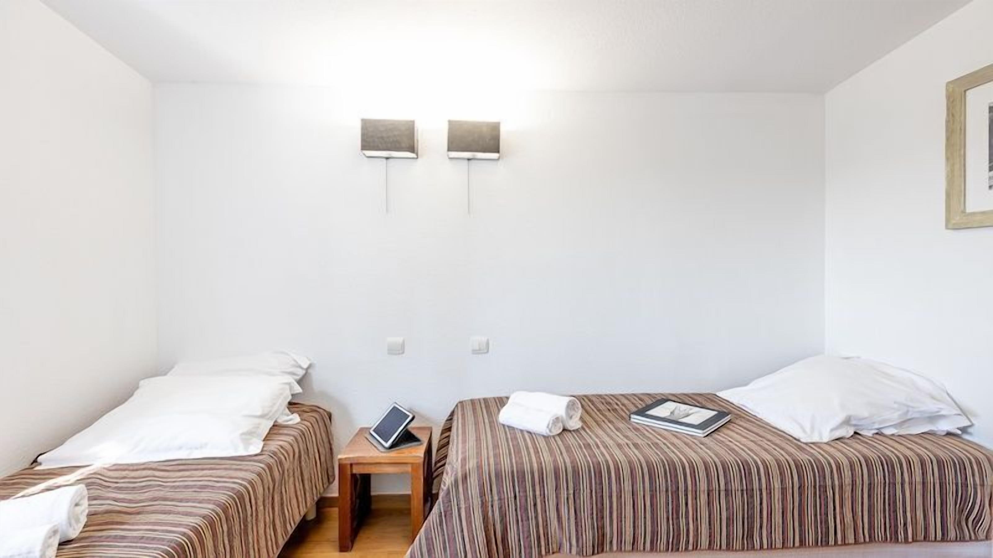 Une petite chambre propre, avec deux lits, une table de chevet et un luminaire fixé au mur.