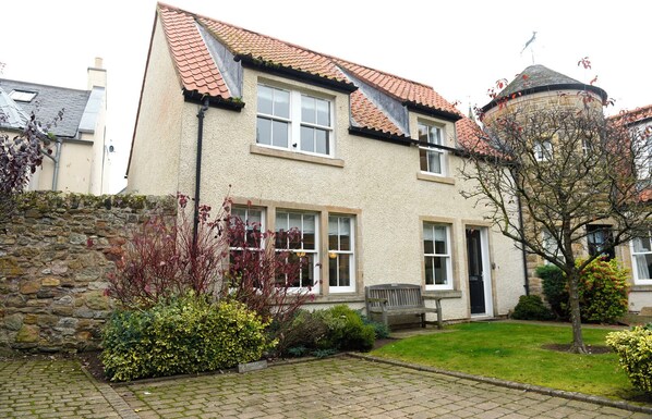 A two-story house with a stone wall, a tiled roof, and a garden with a bench.