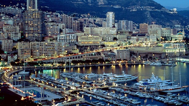 A marina with numerous boats docked, surrounded by a cityscape with high-rise buildings and a mountainous backdrop. - A Roof Between the Sky and the Sea (Sanremo)