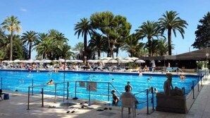 A swimming pool surrounded by palm trees and umbrellas, with people swimming and relaxing. - A Roof Between the Sky and the Sea (Sanremo)