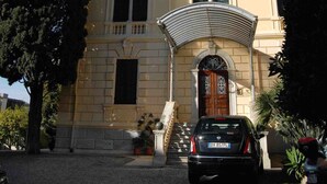 A two-story building with a classic architectural style, featuring a central entrance with a decorative archway and a staircase leading to the upper floor. - A Roof Between the Sky and the Sea (Sanremo)