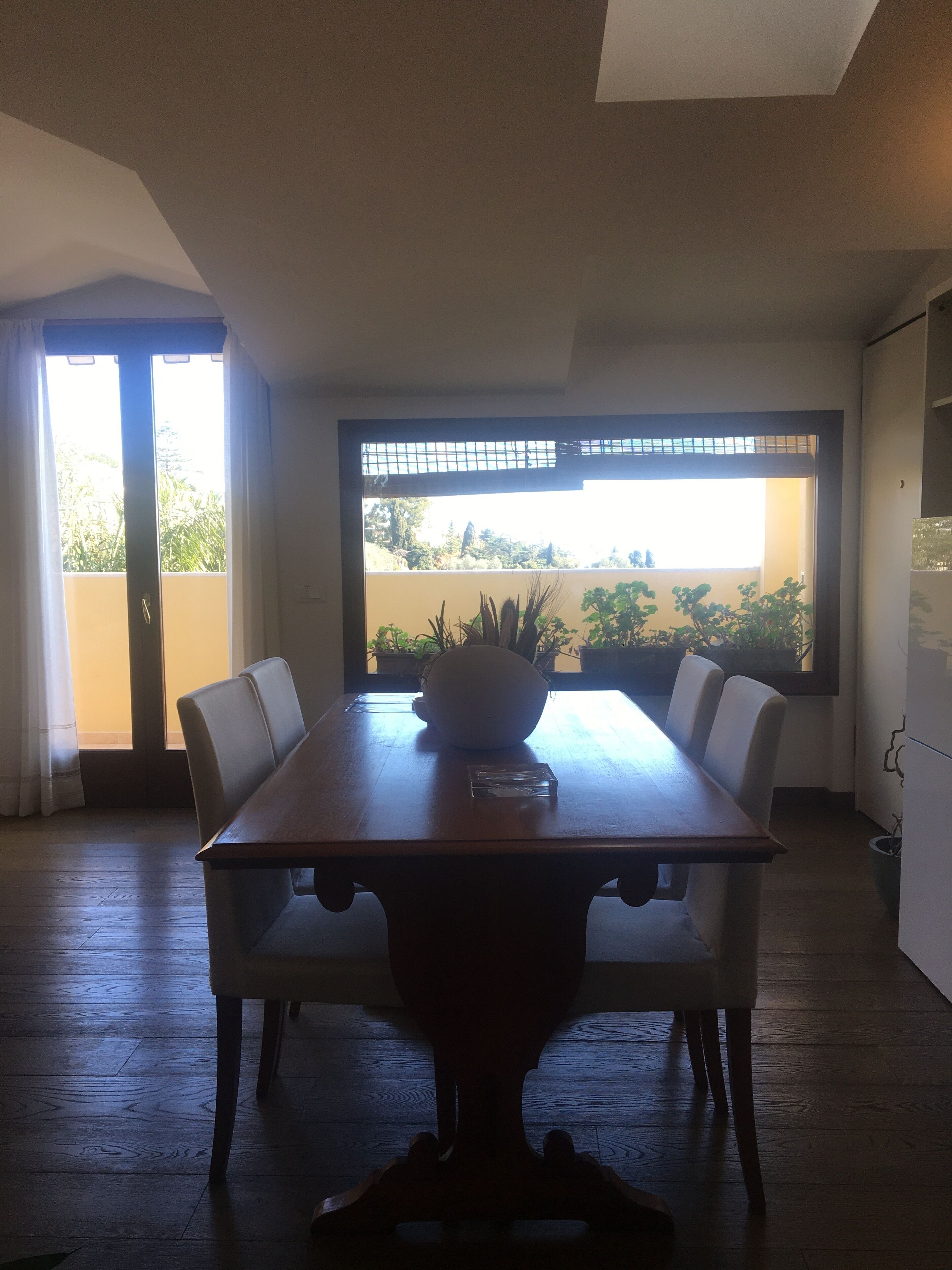 A dining area with a wooden table, white chairs, and a bowl on the table. There is a window with a view of greenery outside.