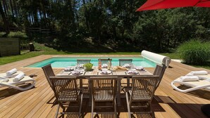 A wooden deck with a pool table set up for dining, surrounded by lounge chairs and a red umbrella.