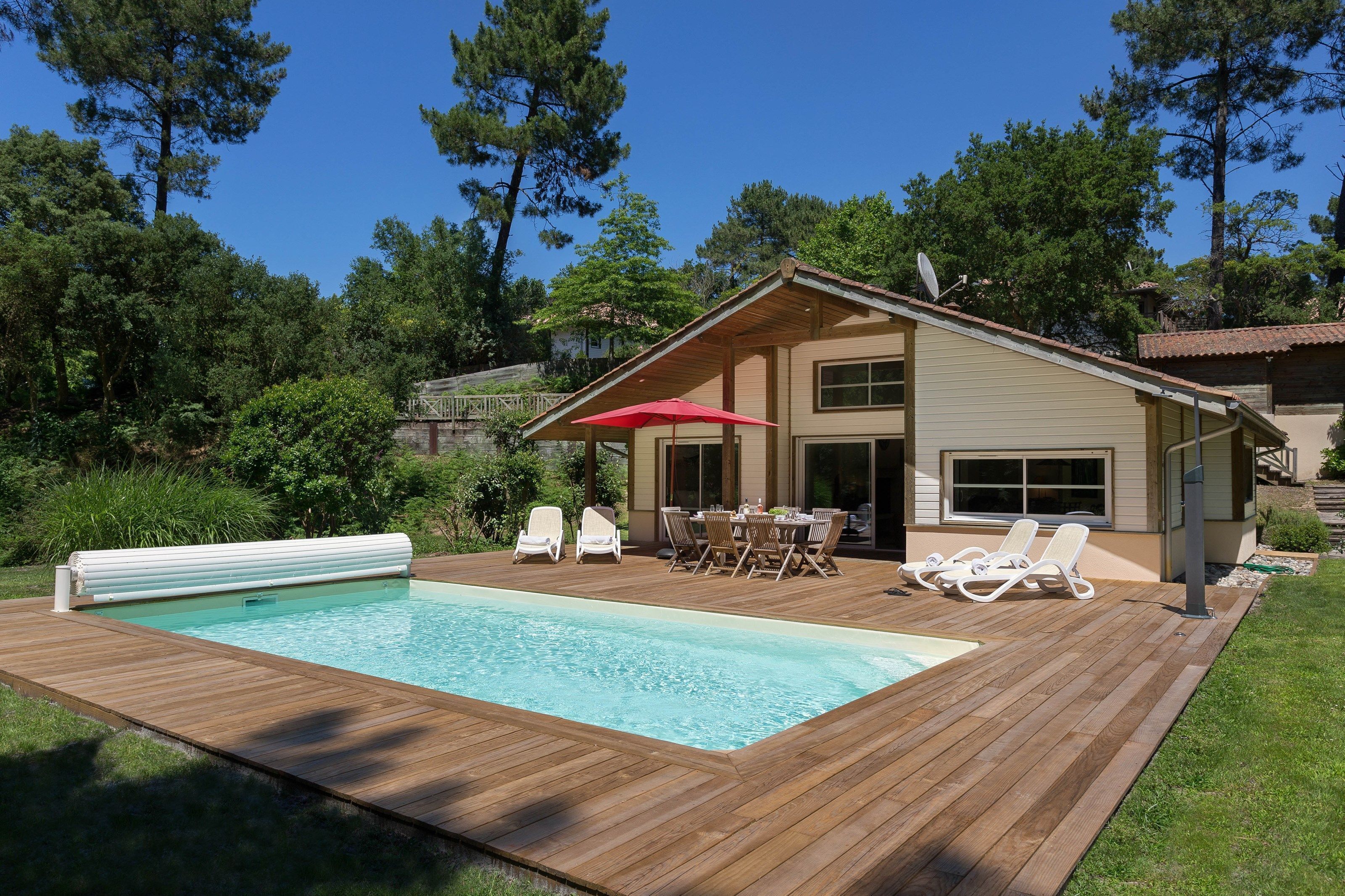 Una terraza de madera con piscina, un porche cubierto con mesa y sillas, y una pequeña construcción con antena parabólica.