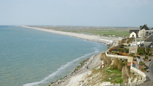 Une vue côtière comprenant une plage, une route menant à une zone située au pied d’une falaise, et des bâtiments à l’architecture traditionnelle.
