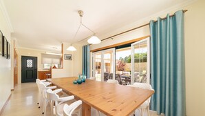 Een ruime eethoek met een houten tafel en witte stoelen, een keuken op de achtergrond, en uitzicht door grote glazen deuren.