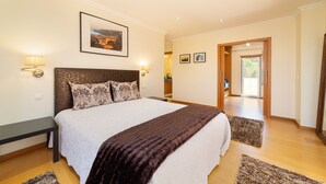 Een ruime slaapkamer met een groot bed, een nachtkastje en een bijzettafeltje. Er hangt een ingelijst beeld aan de muur en een deur die leidt naar een andere kamer.