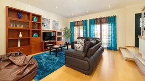 Een moderne woonkamer met een bruine leren bank, houten planken met decoratieve objecten, een flatscreen televisie en een blauw vloerkleed.