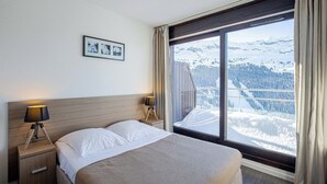 A hotel room with a bed, two bedside lamps, a framed picture on the wall, and a large window with a snowy mountain view. - Appart au pied du téléphérique ! Jacuzzi sur place (FLAINE)