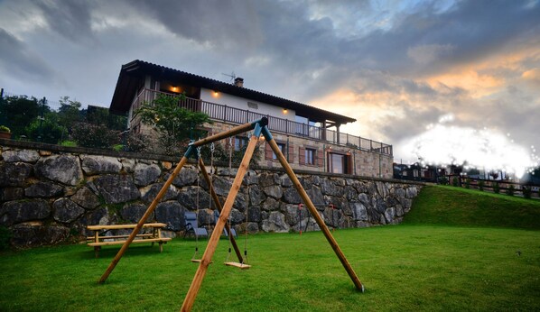 Un conjunto de columpios de madera en un área con césped, una pared de piedra y un edificio al fondo.
