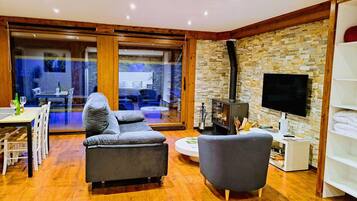 Een moderne woonkamer met een bank, fauteuil en een televisie. Een open haard en een eethoek zijn zichtbaar door een glazen deur.